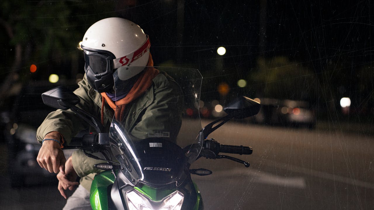 A person on a motorcycle at night, wearing a protective helmet and jacket, on an urban street.
