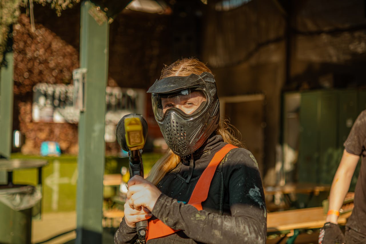 Young woman wearing protective paintball gear and mask posing with a paintball gun outdoors.