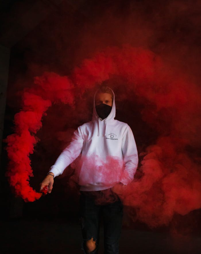 Man in white hoodie with face mask surrounded by swirling red smoke indoors.